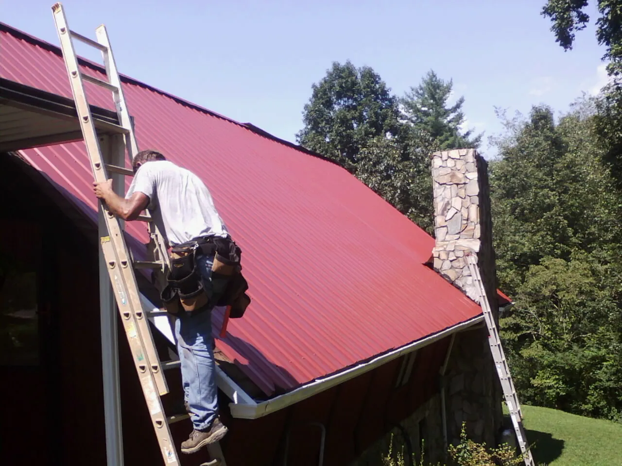 Roofer on a ladder beside a red metal roof and stone chimney — Soffit & Fascia Repair by Roofing Brothers in South Glastonbury